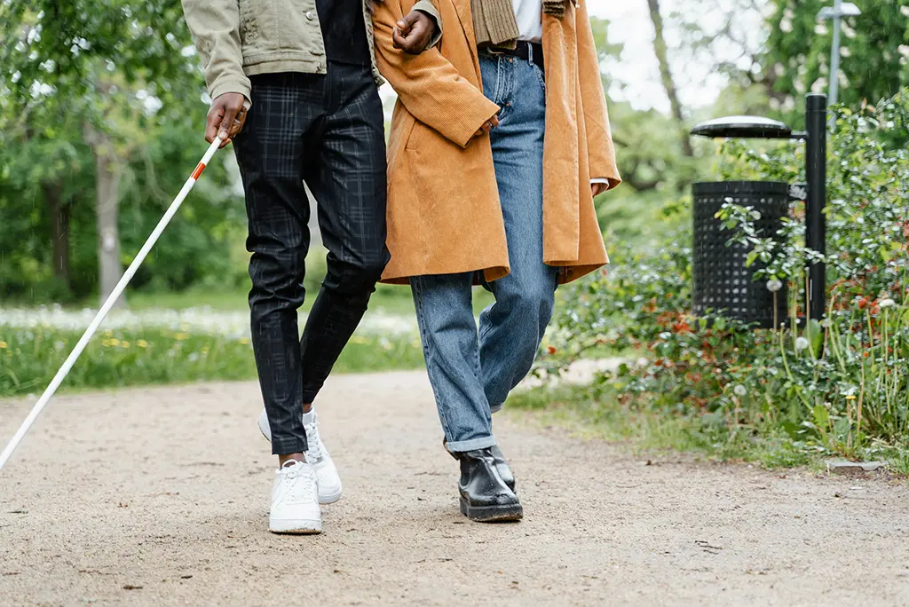 Services for visually impaired adults showing a blind man with cane walking with a female friend through the park.