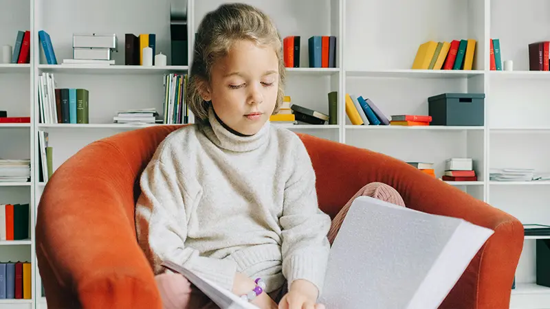 White bookcase behind girl sitting in orange chair with vision loss holding a large book