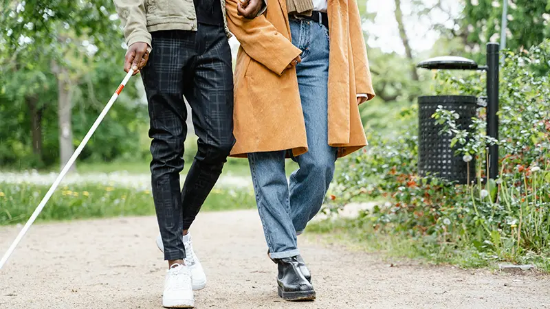 Services for people with vision loss showing a blind man with cane walking with a female friend through the park.